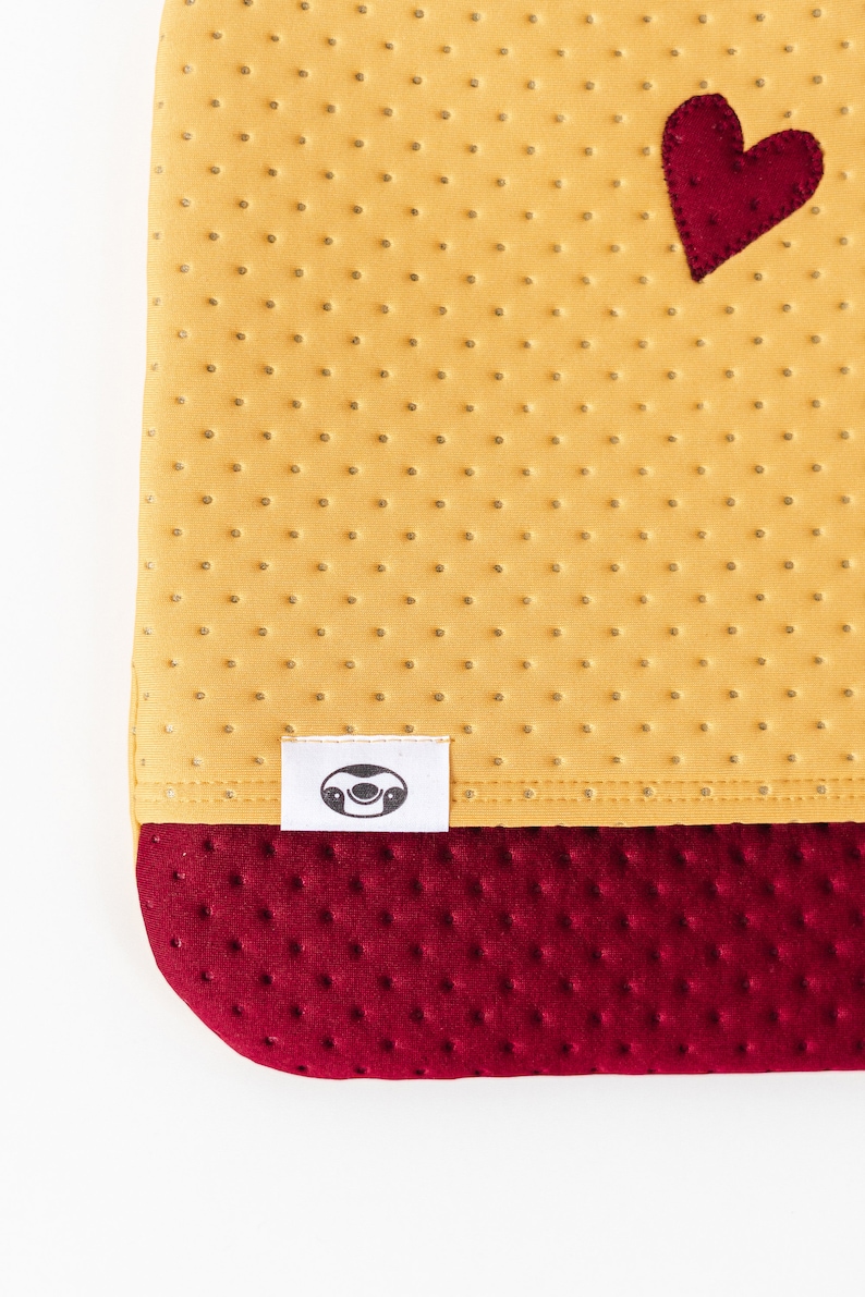 May include: A yellow fabric with a red heart and a white label with a sloth face. The fabric has a textured pattern of small dots. The bottom of the fabric is a darker red color with a similar textured pattern.