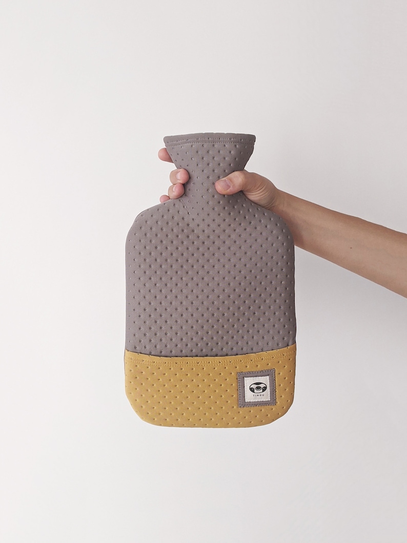 Puede incluir: Una botella de agua caliente gris y amarilla con una cubierta de tela texturizada. La botella tiene un peque&ntilde;o logotipo con la palabra "Tinu" en ella.