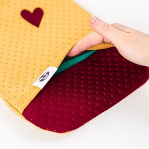 May include: A yellow and burgundy hot water bottle with a heart on the front. The bottle is made of a soft, textured material and has a green lining.