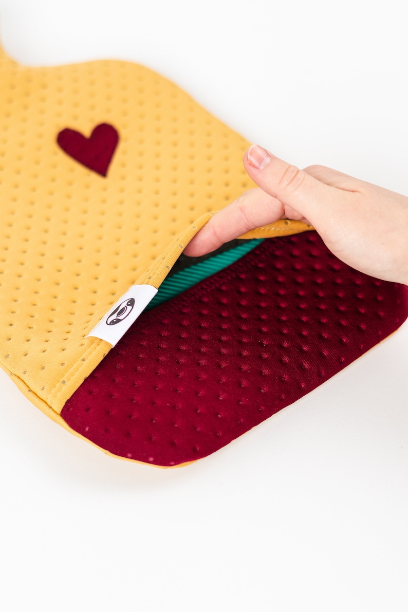 May include: A yellow and burgundy hot water bottle with a heart on the front. The bottle is made of a soft, textured material and has a green lining.