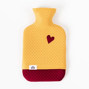 May include: A yellow hot water bottle with a red heart on the front and a red quilted bottom. The bottle has a small white label with a logo on it.
