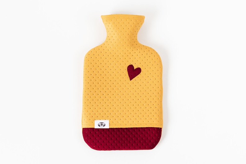 May include: A yellow hot water bottle with a red heart on the front and a red quilted bottom. The bottle has a small white label with a logo on it.