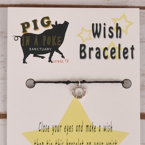 Puede incluir: Un brazalete de cuerda negro con un dije de cerdo plateado. La tarjeta dice "PIG IN A POKE SANCTUARY Lorena, TX" y "Wish Bracelet". La tarjeta también incluye instrucciones: "Cierra los ojos y pide un deseo, luego ata este brazalete en tu muñeca. Cuando se caiga o el hilo se desgaste, ¡tu deseo se hará realidad!"