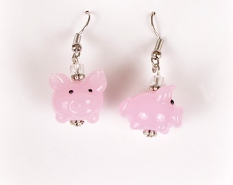 May include: A pair of pink pig earrings with silver hooks and beads. The earrings are on a white background with a logo that says "Pig in a Poke Sanctuary, Lorena, TX".