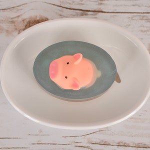 May include: A pink pig shaped soap bar in a clear oval soap dish with a light blue swirl. The soap dish is sitting on a white surface.