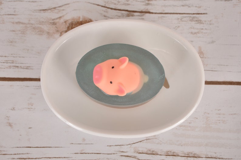 May include: A pink pig shaped soap bar in a clear oval soap dish with a light blue swirl. The soap dish is sitting on a white surface.