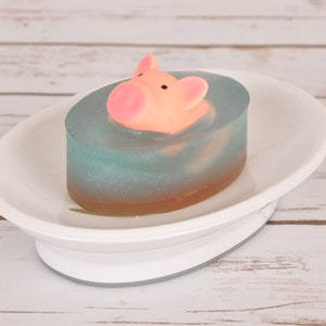 May include: A blue and brown soap bar with a pink pig figurine on top. The soap is sitting on a white oval soap dish.