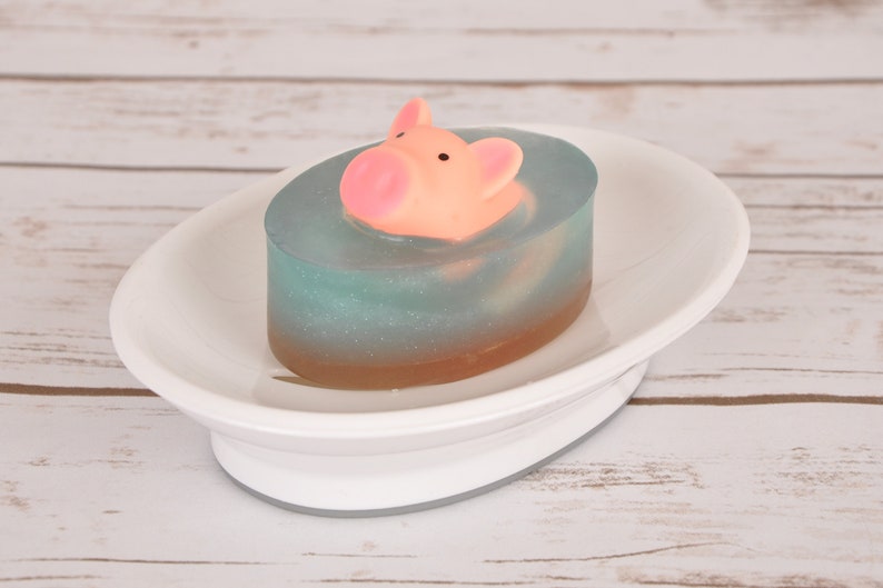 May include: A blue and brown soap bar with a pink pig figurine on top. The soap is sitting on a white oval soap dish.