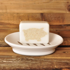 May include: A white bar of soap with a pig design on a white soap dish. The soap dish is on a wooden surface.
