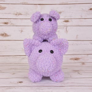 Puede incluir: Dos juguetes de peluche de cerdo lavanda hechos a mano, uno apilado encima del otro. Los cerdos tienen ojos de botón negros y están hechos de un hilo texturizado con motas de colores. El fondo es un veteado de madera clara.