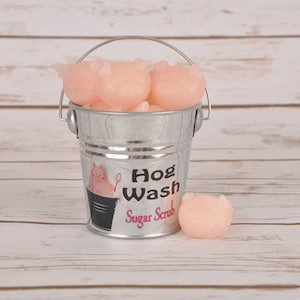 May include: A small metal bucket filled with pink pig-shaped sugar scrub bars. The bucket has a black and white label that reads "Hog Wash Sugar Scrub".