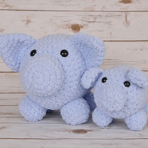 Puede incluir: Dos juguetes de peluche de cerdo hechos a crochet, uno más grande que el otro, ambos de color azul claro. Están sentados sobre una superficie de madera clara.