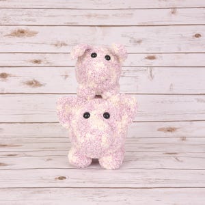 May include: Two handmade, plush pig toys in shades of lavender and white. The larger pig is at the bottom, with a smaller pig stacked on top. Both have black button eyes. The toys are made of a soft, textured material.