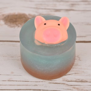 May include: A round, light blue soap with a pink pig head on top. The soap has a shimmery, glittery finish and a brown base.