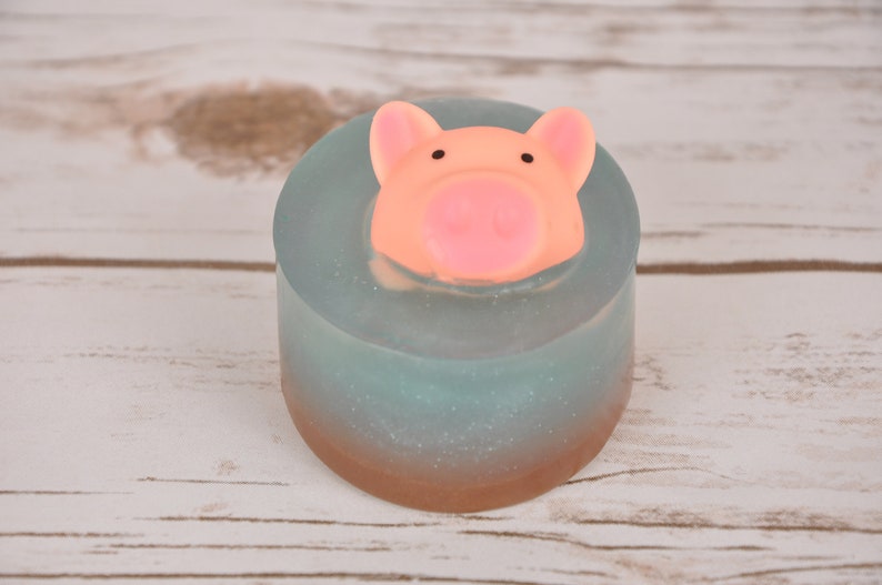 May include: A round, light blue soap with a pink pig head on top. The soap has a shimmery, glittery finish and a brown base.