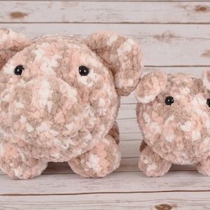 Puede incluir: Dos peluches de cerdo de crochet, uno más grande que el otro, ambos con hilo rosa y marrón y ojos negros. Los peluches están sentados sobre una superficie de madera clara.