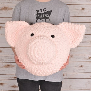 Puede incluir: Una persona sosteniendo una gran cabeza de cerdo de crochet rosa. El cerdo tiene una nariz negra y ojos negros.