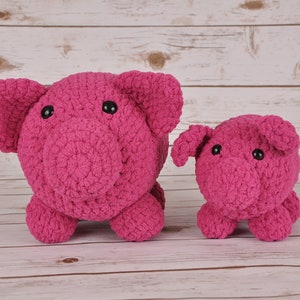 Puede incluir: Dos peluches de cerdo de crochet rosa, uno más grande que el otro, sobre un fondo de madera blanca.