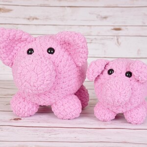 Puede incluir: Dos peluches de cerdo de crochet rosa, uno más grande que el otro, con ojos de botón negros. Los peluches están sentados sobre una superficie de madera blanca.