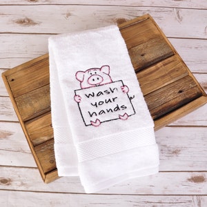 May include: White hand towel with an embroidered pig holding a sign that reads "Wash your hands." The pig is pink with black outlines. The towel is folded and has a textured border.