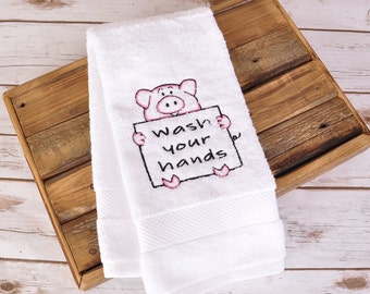 May include: White hand towel with an embroidered pig holding a sign that reads "Wash your hands." The pig is pink with black outlines. The towel is folded and has a textured border.