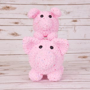 Puede incluir: Dos juguetes de cerdos rosas de peluche apilados uno encima del otro. El cerdo inferior tiene orejas grandes y un cuerpo redondo. El cerdo superior tiene un cuerpo y orejas más pequeños. Ambos tienen ojos de botón negros.