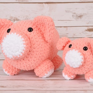 Puede incluir: Dos peluches de cerdo de crochet, uno más grande que el otro, ambos de color rosa claro con hocicos blancos y ojos negros. Los peluches están sentados sobre una superficie de madera clara.