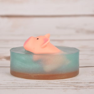 May include: A light blue and brown soap bar with a pink pig figurine floating inside. The soap bar is on a white wooden surface.