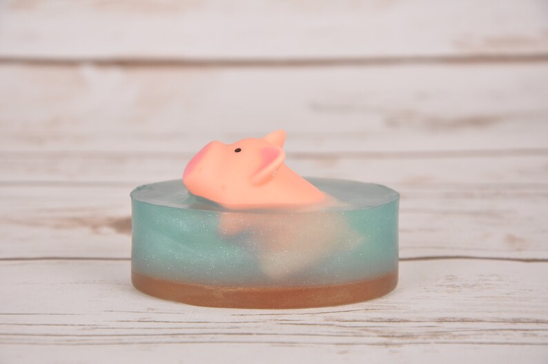 May include: A light blue and brown soap bar with a pink pig figurine floating inside. The soap bar is on a white wooden surface.