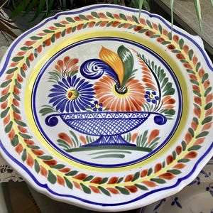 Könnte beinhalten: Ein dekorativer Keramikteller mit gewelltem Rand. Der Teller zeigt ein zentrales Blumendesign in Blau, Orange und Gelb, das in einem blauen Korb angeordnet ist. Ein Rand aus grünen, orangefarbenen und gelben Blättern umgibt den Teller.