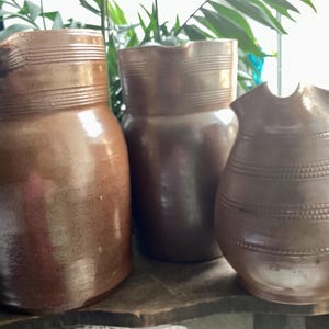 May include: Three brown ceramic pitchers of varying sizes. The pitchers have a rustic, handmade appearance with horizontal banded detailing. The largest pitcher has a spout. The pitchers are displayed on a wooden surface.