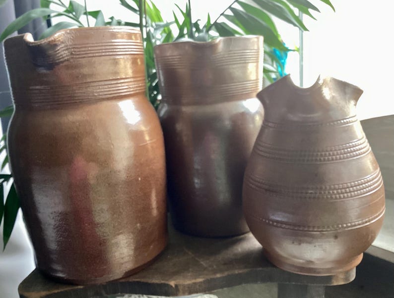 May include: Three brown ceramic pitchers of varying sizes. The pitchers have a rustic, handmade appearance with horizontal banded detailing. The largest pitcher has a spout. The pitchers are displayed on a wooden surface.