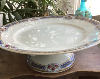 Puede incluir: Soporte para pasteles de cerámica blanca con un plato ancho y poco profundo y una base de pedestal. El borde y la base están decorados con un borde azul y dorado y detalles florales. Para exhibir pasteles o postres.