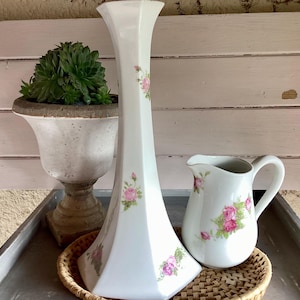 May include: A tall white hexagonal porcelain vase and a small white porcelain creamer pitcher, both decorated with delicate pink rose floral patterns and green leaves, arranged together on a round woven wicker tray surface.