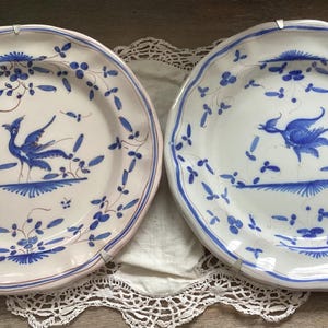 May include: Two white ceramic plates with blue floral and bird designs. The plates have scalloped edges and are decorated with blue birds and floral patterns. The plates are displayed on a white lace doily.