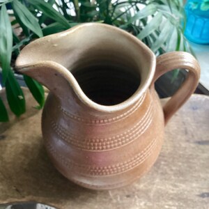 May include: A brown ceramic pitcher with a curved spout and a handle. The pitcher has horizontal bands of dotted texture around its body. The pitcher is likely used for serving beverages and has a rustic, handmade appearance.