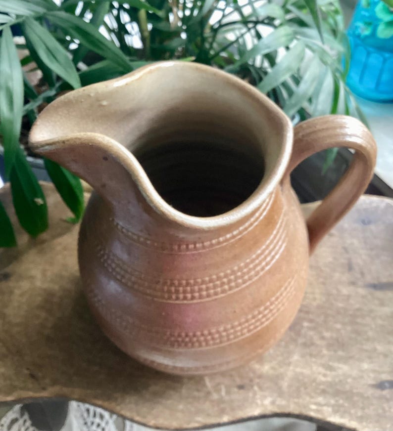 May include: A brown ceramic pitcher with a curved spout and a handle. The pitcher has horizontal bands of dotted texture around its body. The pitcher is likely used for serving beverages and has a rustic, handmade appearance.