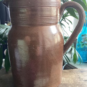 May include: A brown ceramic pitcher with a handle. The pitcher has a wide base and a narrower top with a spout. The top of the pitcher has horizontal ridges. The handle is curved and textured. The pitcher is likely used for serving beverages.
