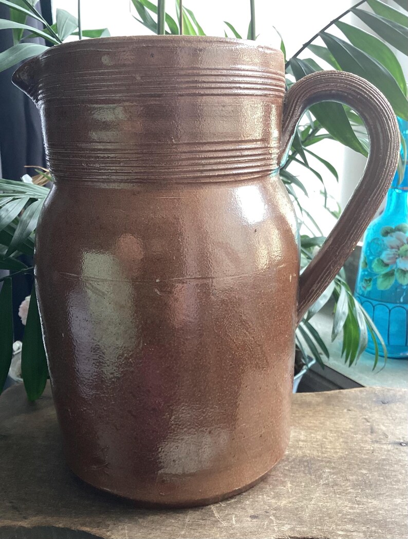 May include: A brown ceramic pitcher with a handle. The pitcher has a wide base and a narrower top with a spout. The top of the pitcher has horizontal ridges. The handle is curved and textured. The pitcher is likely used for serving beverages.