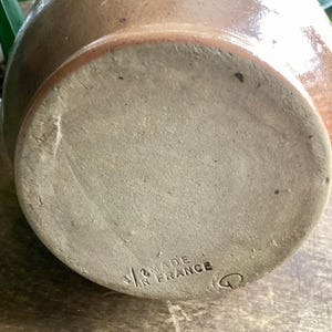 May include: A close-up of the bottom of a brown ceramic jug. The base is a lighter tan color with the words "MADE IN FRANCE" stamped on it. The jug has a rounded shape and a slightly textured surface.