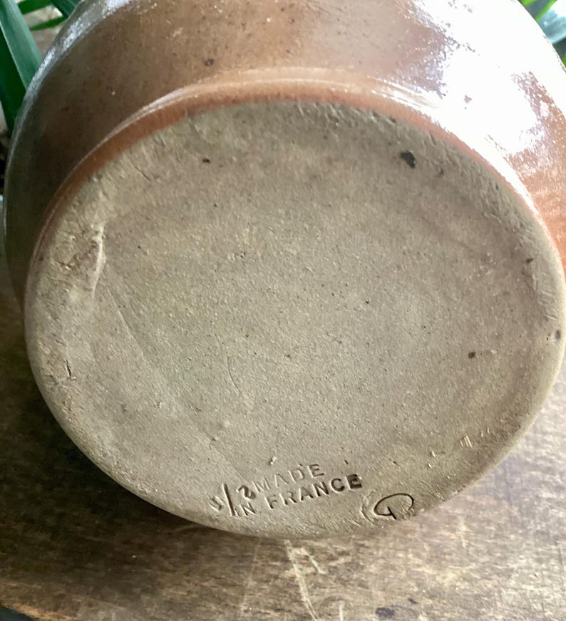 May include: A close-up of the bottom of a brown ceramic jug. The base is a lighter tan color with the words "MADE IN FRANCE" stamped on it. The jug has a rounded shape and a slightly textured surface.