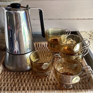 May include: A polished stainless steel stovetop coffee maker with a black handle and lid knob, accompanied by four amber-coloured glass cups featuring a textured base and a small circular gold-toned sticker on the side of each glass vessel.