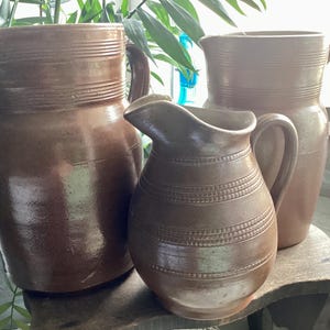 May include: Three brown ceramic pitchers of varying sizes. The largest pitcher has a wide base and a handle. The middle pitcher is smaller with a rounded body and a spout. The smallest pitcher is in the front. All have horizontal banded details.