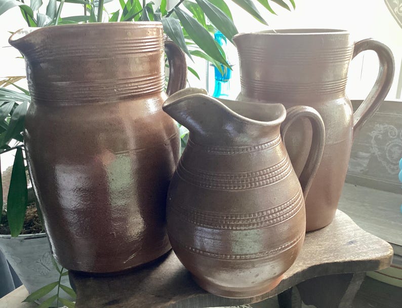May include: Three brown ceramic pitchers of varying sizes. The largest pitcher has a wide base and a handle. The middle pitcher is smaller with a rounded body and a spout. The smallest pitcher is in the front. All have horizontal banded details.
