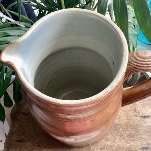 May include: A handmade, brown ceramic pitcher with a wide opening and a handle. The interior is a light blue-gray color. The pitcher has a spout for easy pouring and is likely used for serving beverages.