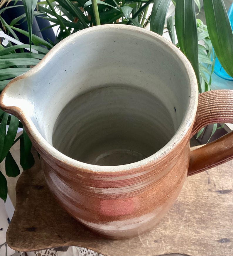 May include: A handmade, brown ceramic pitcher with a wide opening and a handle. The interior is a light blue-gray color. The pitcher has a spout for easy pouring and is likely used for serving beverages.