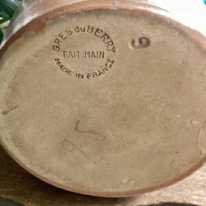 May include: Close-up of the bottom of a brown stoneware pottery piece. The text "GRES du BERRY FAIT MAIN MADE IN FRANCE" is stamped in a circle. A number "9" and other markings are also visible.