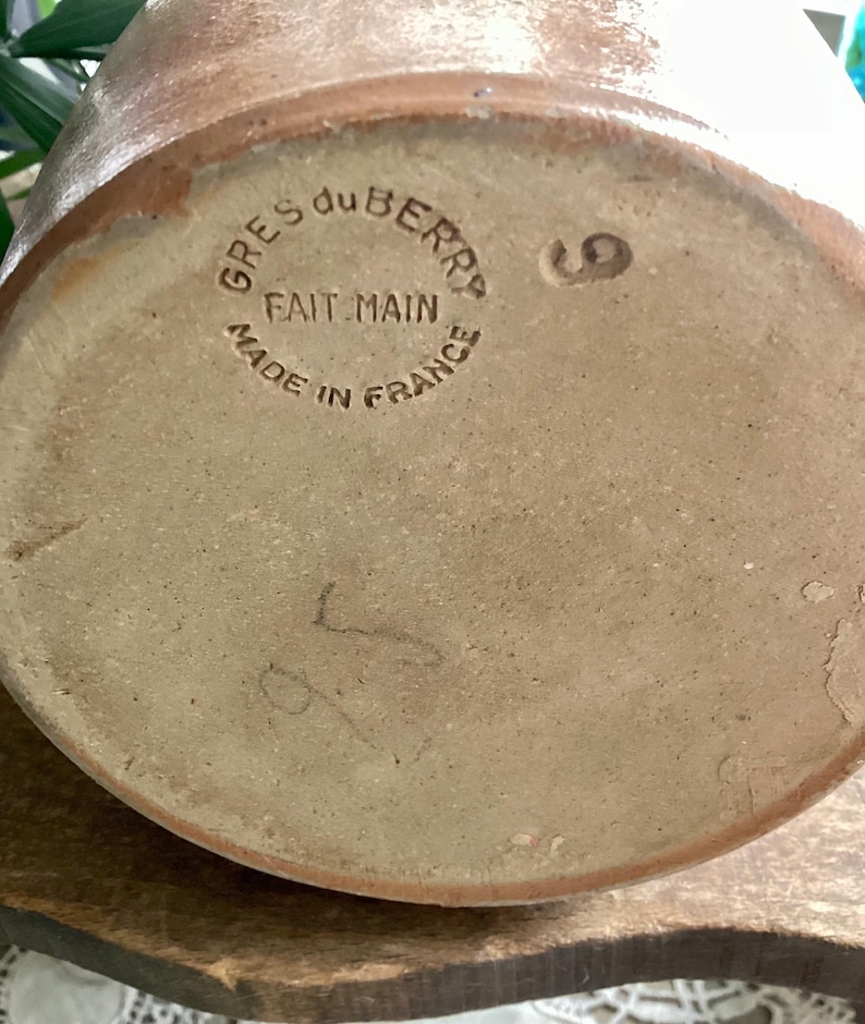 May include: Close-up of the bottom of a brown stoneware pottery piece. The text "GRES du BERRY FAIT MAIN MADE IN FRANCE" is stamped in a circle. A number "9" and other markings are also visible.