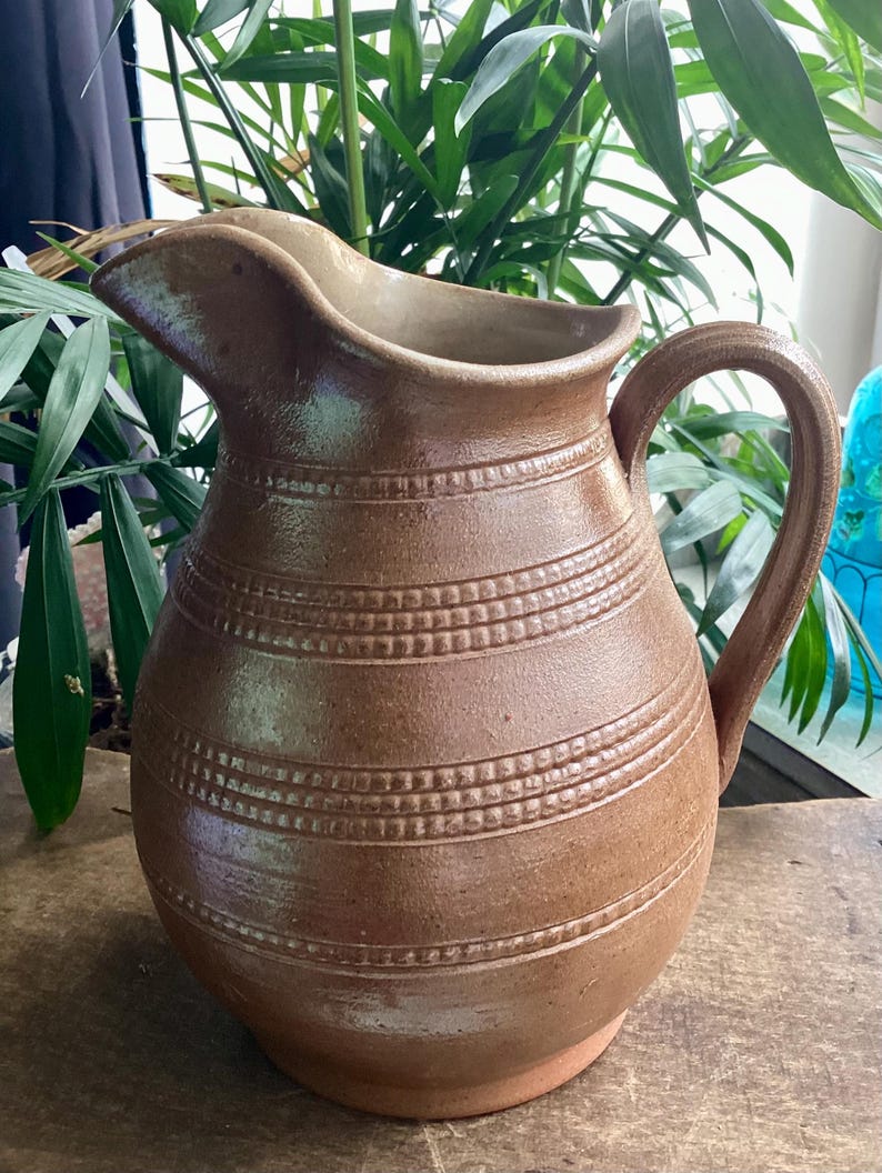 May include: A brown ceramic pitcher with a handle and spout. The pitcher has horizontal bands of textured detailing around the body. The pitcher is sitting on a wooden surface, with green plants in the background. The pitcher is likely used for serving beverages.