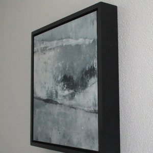 Op de afbeelding: Abstract kunstwerk in een zwarte lijst. Het kunstwerk toont een gestructureerd, gelaagd ontwerp in grijstinten, wit en zwart. Het schilderij heeft een moderne esthetiek, met zichtbare penseelstreken en een minimalistische stijl. De lijst is vierkant en heeft een matte afwerking.
